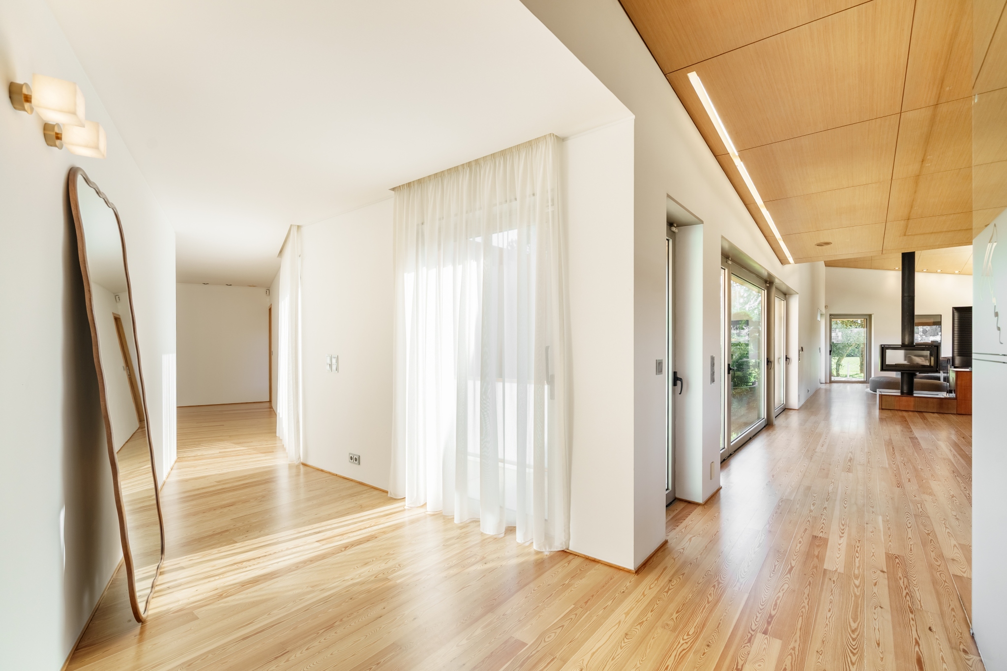 An open living space — pine floor, white walls, sheer curtains and light from every side.
