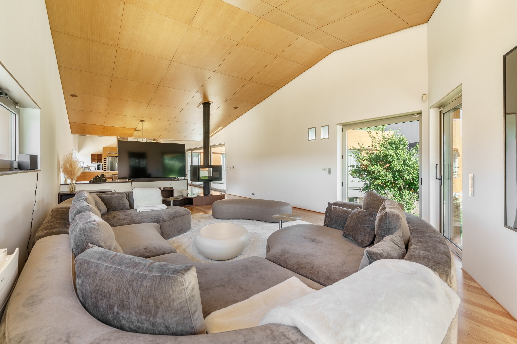 Open living room with curved sofa, wood-burning stove and floor-to-ceiling glass to the garden.